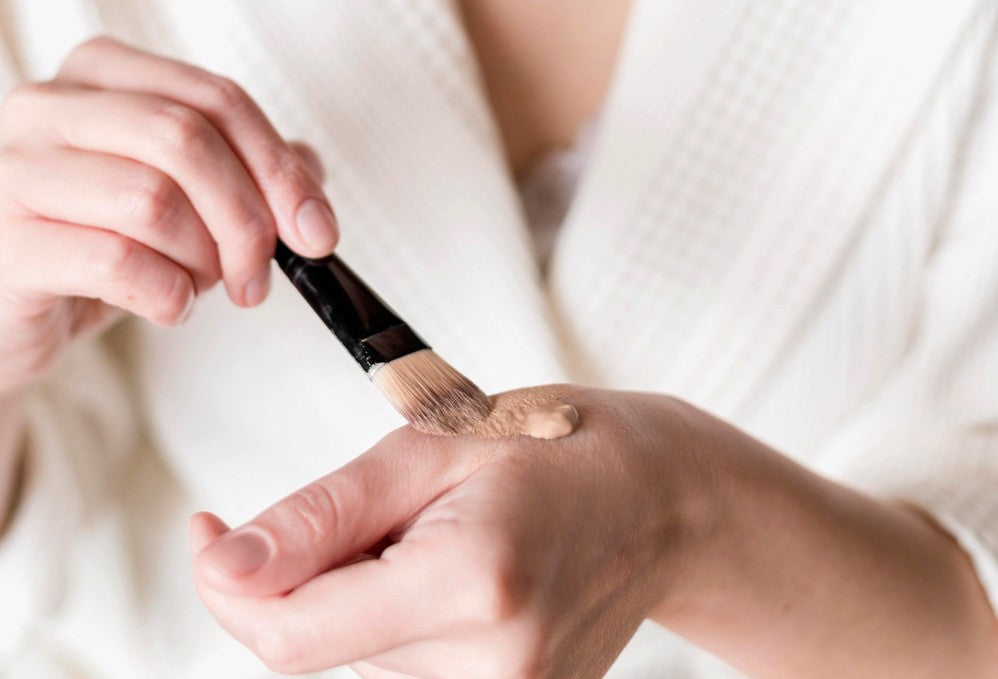 Primer plano de la mano de una mujer que tiene en el dorso base de maquillaje que toma con una brocha para aplicarlo en su cara.
