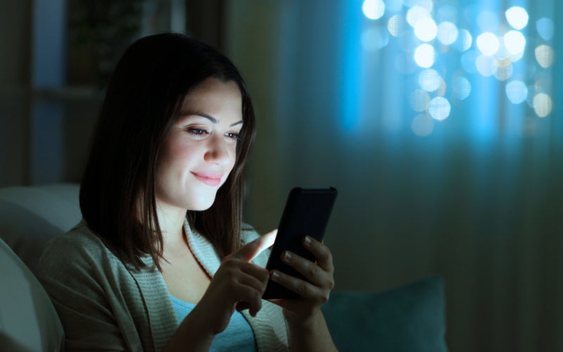 Mujer joven mirando su celular de noche mostrando la exposición diaria a la luz azul y su impacto en la piel