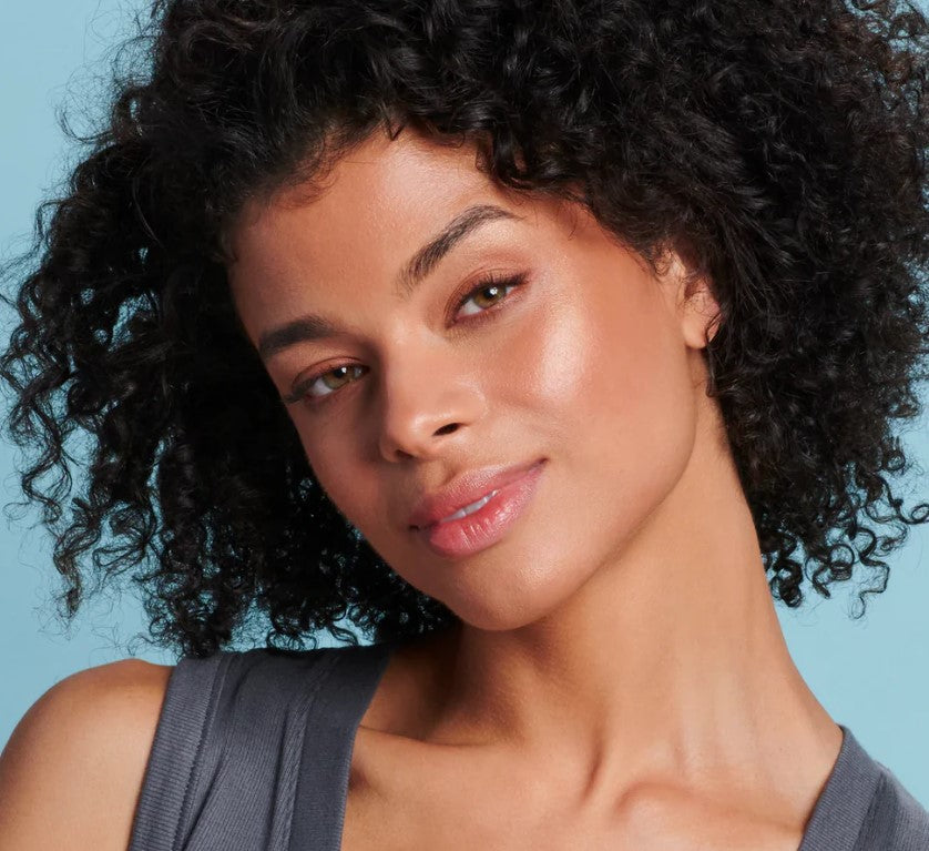 una mujer con cabello rizado oscuro y piel bronceada, luce un maquillaje natural que la hace ver saludable y natural, ya que resalta sus mejillas y el puente de su nariz. Sus labios tienen un color sutil.