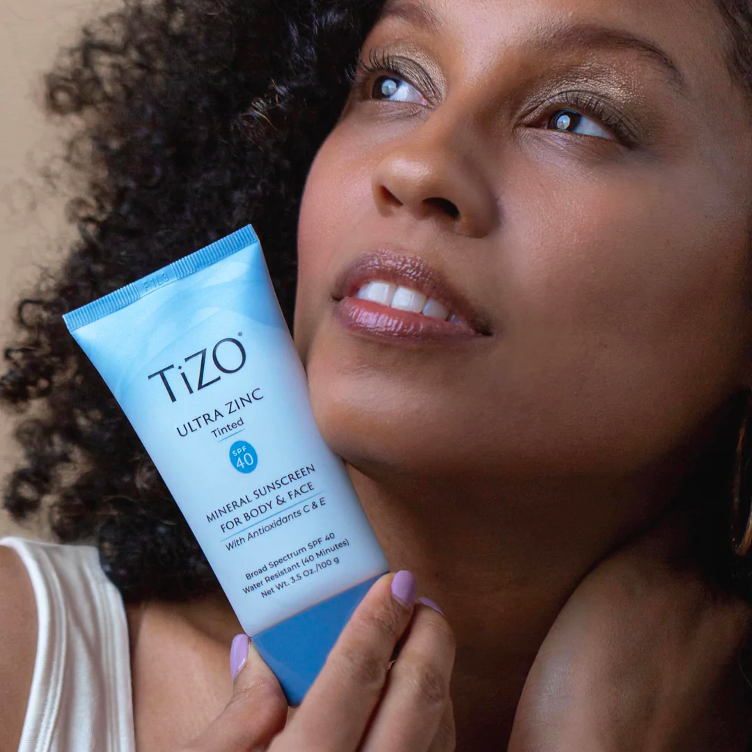 Mujer de piel morena y cabello rizado sosteniendo Tizo Ultra Zinc SPF 40 con color cerca del rostro, en fondo neutro.