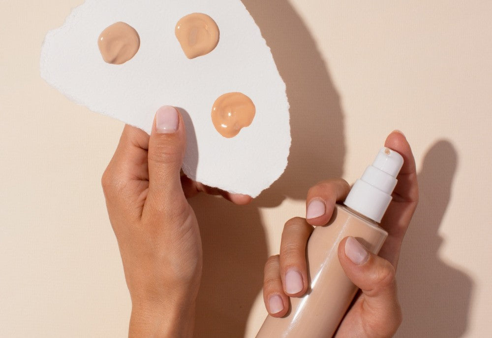Vista de una mano que sostiene un trozo de papel donde hay tres muestras de base de maquillaje líquida, de diferente tono. La otra sostiene un envase con el producto. 