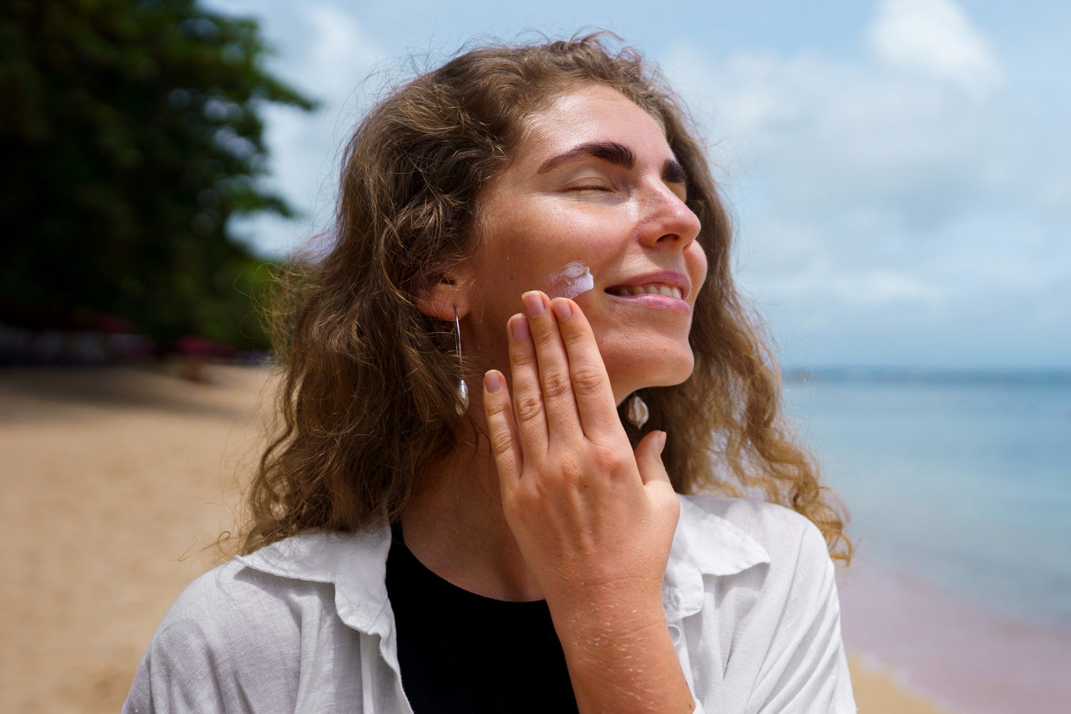 Mujer sonriente, con cabello rizado, aplica protector solar en su rostro. De fondo se ve un paisaje en la playa. La imagen ilustra el uso de un fotoprotector físico.