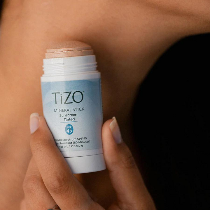 Persona aplicando TiZO Mineral Stick Tinted SPF 45 en la piel del cuello, mostrando el envase abierto en primer plano.