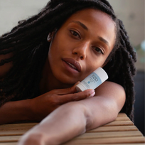 Mujer recostada mirando a cámara mientras sostiene el envase del protector solar TiZO Mineral Stick Tinted SPF 45 sobre su brazo extendido.